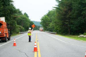 A flagger sends traffic up Sylvan Road to Grafton Road to go around the repair work. The oversize load can be seen in the distance.
